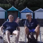 Two man sit in chairs on the beach talking to each other in a still from the 2024 film Red Herring